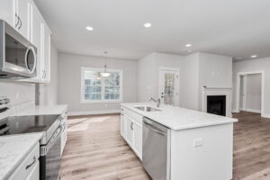 A bright, modern kitchen with white cabinets, marble countertops, and hardwood floors, featuring a central island and a fireplace in the adjoining room.