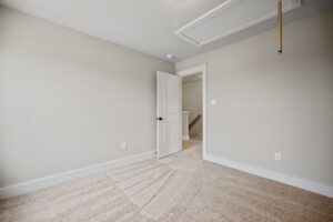 Empty beige carpeted room with an open white door leading to a staircase. A ceiling access panel is visible above the door. Walls and baseboards are white.