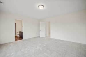 A spacious, empty room with a closed door, an open door leading to a bathroom, beige carpet, and a ceiling light fixture. The bathroom has a visible toilet and a wooden vanity.