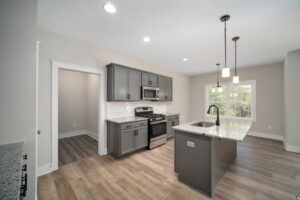A modern kitchen with grey cabinets, white subway tile backsplash, stainless steel appliances, an island with a granite countertop, pendant lighting, and hardwood flooring.