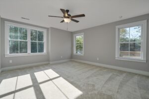 A bright, empty room with beige carpet, gray walls, a ceiling fan with a light, and three large windows letting in natural light.