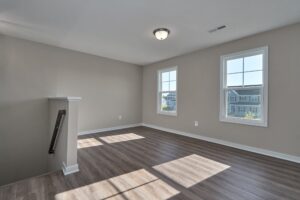An empty room with light gray walls, two windows, and wood-look flooring; a small staircase is partially visible on the left.