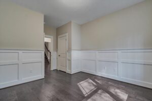 An empty room with beige walls, white wainscoting, dark wood floors, and a closed door. An open doorway shows a view of stairs in the background. Natural light illuminates part of the floor.