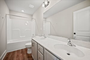 A clean bathroom with a double sink vanity, white countertop, large mirror, bathtub-shower combo, white walls, and wood flooring.