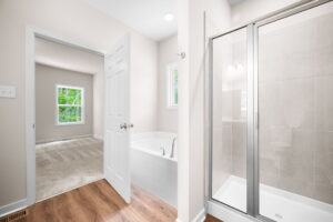 Newly renovated empty bathroom with a glass shower door, beige tiles, adjacent bathtub, and an open doorway leading to a bedroom with a window.