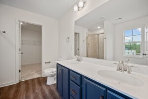 Modern bathroom with a double sink vanity with blue cabinets, large mirror, toilet, and glass-enclosed shower. There is a door to a walk-in closet and a window with an outside view.