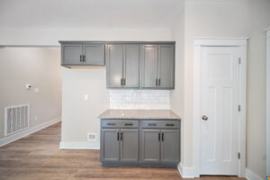 A kitchen area with gray cabinets, granite countertop, white subway tile backsplash, and wooden flooring. There is a white paneled door to the right.