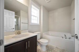 A clean, modern bathroom with a white bathtub, a toilet, a dark wood vanity with a sink and countertop, a large mirror, and a window above the tub.