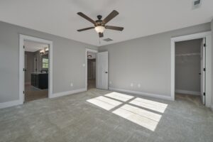 A spacious, carpeted bedroom with light gray walls, a ceiling fan, a walk-in closet on the right, and an open doorway to a bathroom on the left. Natural light enters through a window.