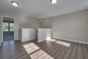 An empty room with wood flooring, beige walls, and two light fixtures on the ceiling. Adjacent is a stairway with a small landing area and a doorway leading to another room.