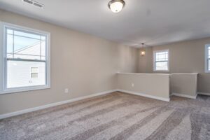 Empty room with beige walls, gray carpet, and three windows. features include a white chair rail and central ceiling light.