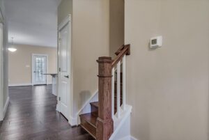 A staircase with wooden handrails and dark steps in a beige-painted interior, leading to a well-lit open hallway with dark wooden floors and a thermostat on the wall.
