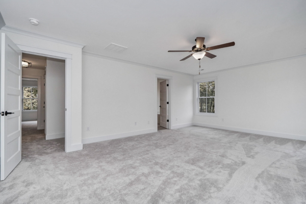 Spacious empty room with light gray carpet, white walls, a ceiling fan, and open doorways leading to other rooms. A window provides natural light.