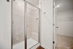 Image of a modern, white-tiled shower with glass doors next to a walk-in closet with wire shelving. The shower features a built-in bench.