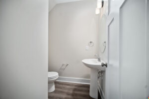 A small white bathroom with a toilet, pedestal sink, circular mirror, and wall-mounted light, featuring light gray walls and dark hardwood flooring.