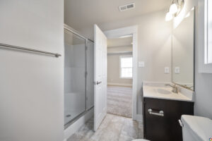A modern bathroom with a glass-enclosed shower, a white toilet, a sink with a dark cabinet, and a large mirror. The bathroom door is open, revealing a carpeted room with a large window.