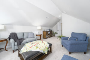 Spacious, well-lit attic living room with slanted ceilings, featuring a patterned ottoman, blue sofas, and matching lamps.