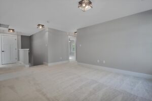 A spacious, carpeted room with grey walls, modern light fixtures, and a white door at the end of a short hallway. Natural light enters from a doorway on the right side.