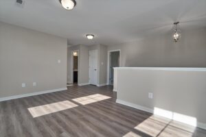 A well-lit empty room with light gray walls, dark wood flooring, and modern light fixtures. The space features a doorway, a half-wall, and several power outlets. Natural light enters from the left.
