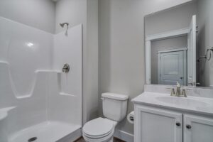 A modern bathroom featuring a white shower and bathtub combo, a toilet, and a vanity with a sink and mirror. The walls are light gray, and the floor has wood-like tiles.