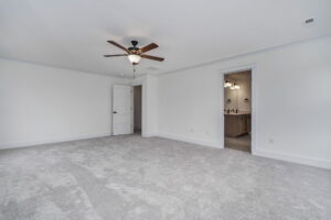 A spacious, carpeted room with a ceiling fan, white walls, a door leading to an adjoining room, and another door leading to a bathroom with a double sink vanity.