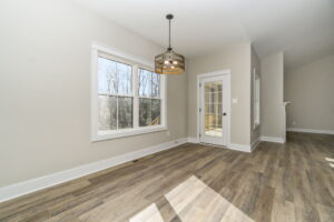 Empty modern living room with large windows, a glass door leading to the outside, and a unique circular chandelier, with light hardwood flooring.