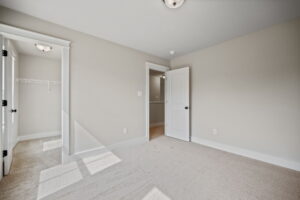 An empty room with beige walls and carpet, lit by natural light from a window on the left. There is an open door to a closet on the left and another open door to a hallway or adjoining room straight ahead.