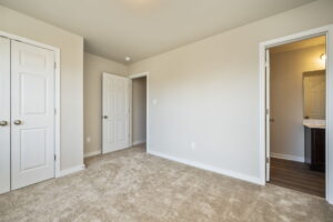 Empty beige carpeted room with white doors and an open door leading to a brightly lit bathroom.
