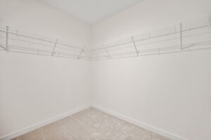 This image shows an empty white room with beige carpet and white wire shelves installed on two walls.