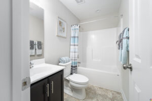 A modern, clean bathroom featuring a white tub with a shower, a toilet, a vanity with a sink, and neatly hung towels. decor includes framed beach-themed art on the walls.