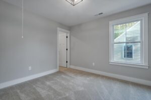 A gray-carpeted room with light gray walls, a window on the right showing part of a house outside, and an open door on the left leading to another room.