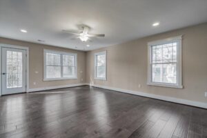 A spacious empty room with hardwood flooring, beige walls, three large windows, a ceiling fan with a light, and a door leading to a balcony.