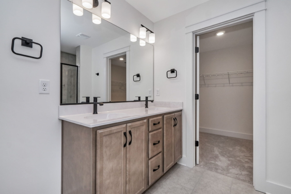 Modern bathroom features a double vanity with two sinks, large mirror, black fixtures, ample cabinet storage, and an adjoining walk-in closet with shelving.