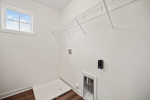 Empty laundry room with a white wire shelf, a dryer vent, and a hook-up for a washing machine. There is a small window on the wall and wooden flooring.