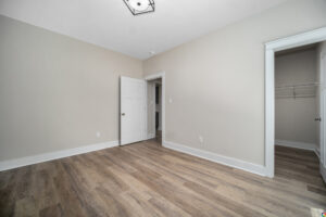 An empty room with light gray walls, a white door, a walk-in closet, and light wood flooring. There is a light fixture on the ceiling.