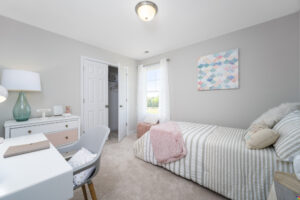 A neatly organized bedroom with a single bed, a study desk, a chair, and a closet. The walls are light-colored, and there is a window with white curtains letting in natural light.