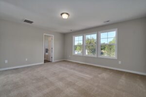 A spacious, empty room with beige carpet, light gray walls, three windows, a ceiling light fixture, and a doorway leading to another room.