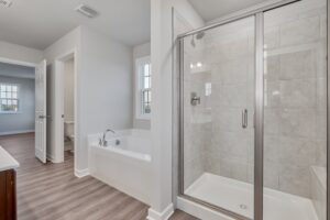 Modern bathroom featuring a glass-enclosed shower, a large bathtub, and a doorway leading to a toilet area. The room has light gray walls and wood flooring.