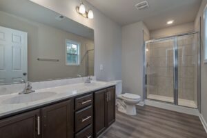 A modern bathroom featuring a double sink vanity with dark cabinets, a toilet, a glass-enclosed shower, and wooden flooring.