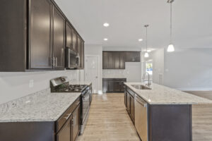 Modern kitchen featuring dark wood cabinets, stainless steel appliances, granite countertops, and a center island with a sink and pendant lighting. Light wood flooring adds warmth to the space.