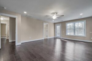 Spacious, unfurnished living room with dark hardwood floors, beige walls, ceiling fan, and several windows. Doorway leads to another room and a door provides access to the outside.