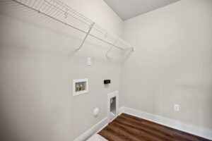 Empty laundry room with a white wire shelf, electrical outlets, utility hook-ups, and a wooden floor. Walls and ceiling are painted light grey.