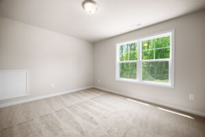 An empty carpeted room with beige walls, a ceiling light, and a double window offering a view of green trees.