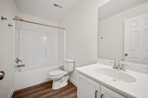 A clean, white bathroom with a bathtub and shower combo, a toilet, a sink with a cabinet, and a large mirror above the sink. The floor is made of light wood.