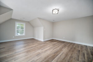 A spacious, empty room with light-colored walls, a window, and wood flooring. It has a modern ceiling light fixture, and angled ceilings on the left side.