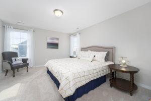 Modern bedroom with a large bed featuring a geometric pattern comforter, decorative pillows, an armchair with a "love" cushion, nightstand with a lamp, and a framed landscape artwork above the bed.