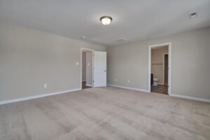 An empty room with beige carpet, light gray walls, a ceiling light fixture, and two doorways, one leading to a bathroom and the other to a hallway.