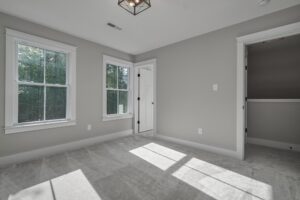 A bright, empty room with grey walls, two windows, a light fixture, and a closed white door. Sunlight is streaming in through the windows, illuminating the room.