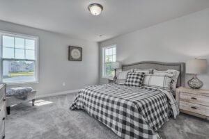 A bedroom with a checkered bedspread, matching pillows, two wooden nightstands with lamps, two windows, and a bench. The walls are light-colored, and the room is carpeted.