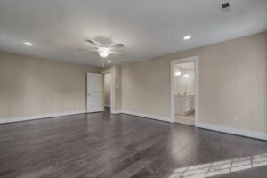A spacious, empty room with hardwood flooring, beige walls, a ceiling fan, recessed lighting, and an open doorway leading to a bathroom with double sinks and mirrors.